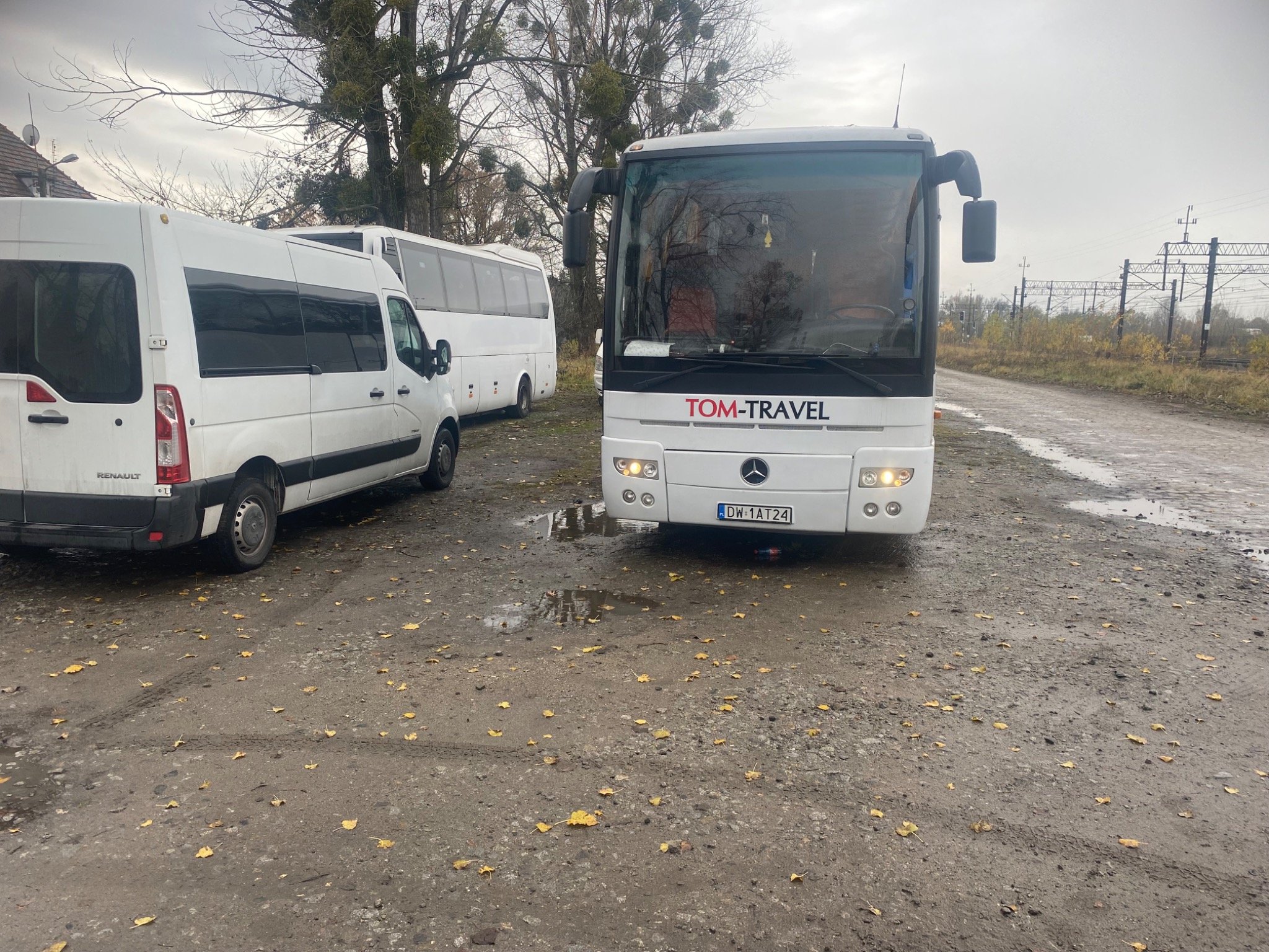 Biały autobus marki Mercedes z logo Tom-Travel parkuje na szarym, lekko zabłoconym placu, obok białego busa Renault i drugiego autobusu w tle, widoczny fragment linii kolejowej.