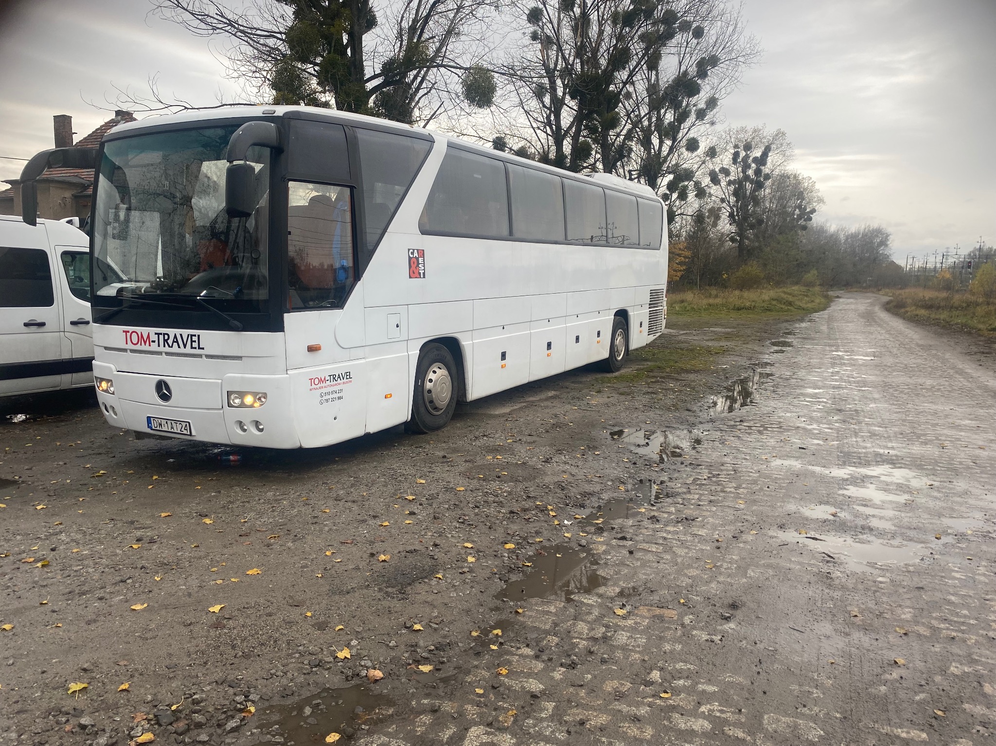 Biały autokar TOM-TRAVEL zaparkowany na błotnistym placu z kałużami i opadłymi liśćmi, w tle drzewa z jemiołą i szara, pochmurna pogoda.