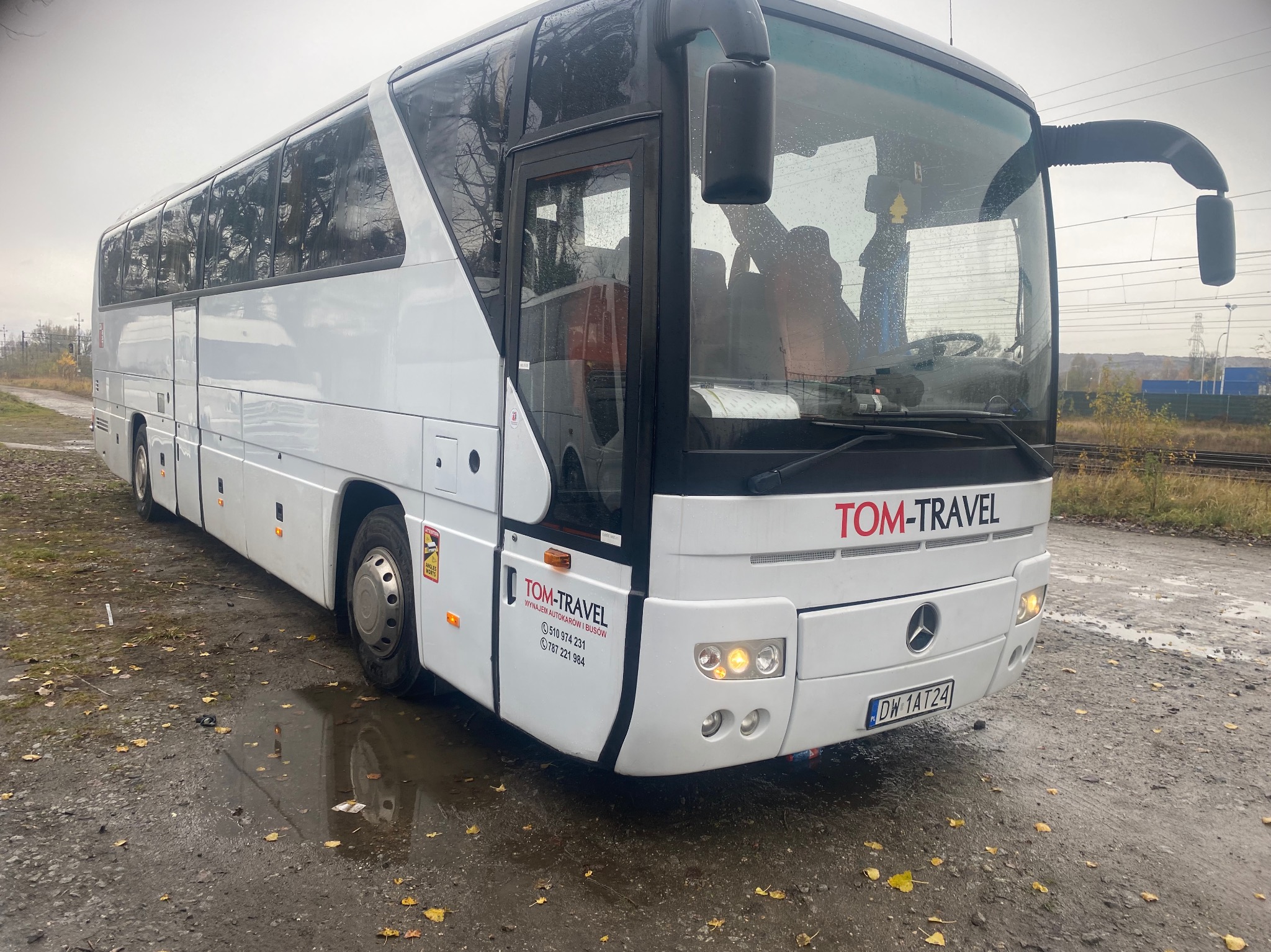 Biały autokar firmy TOM-TRAVEL z widocznymi numerami kontaktowymi na karoserii, stojący na mokrym placu z opadłymi liśćmi.