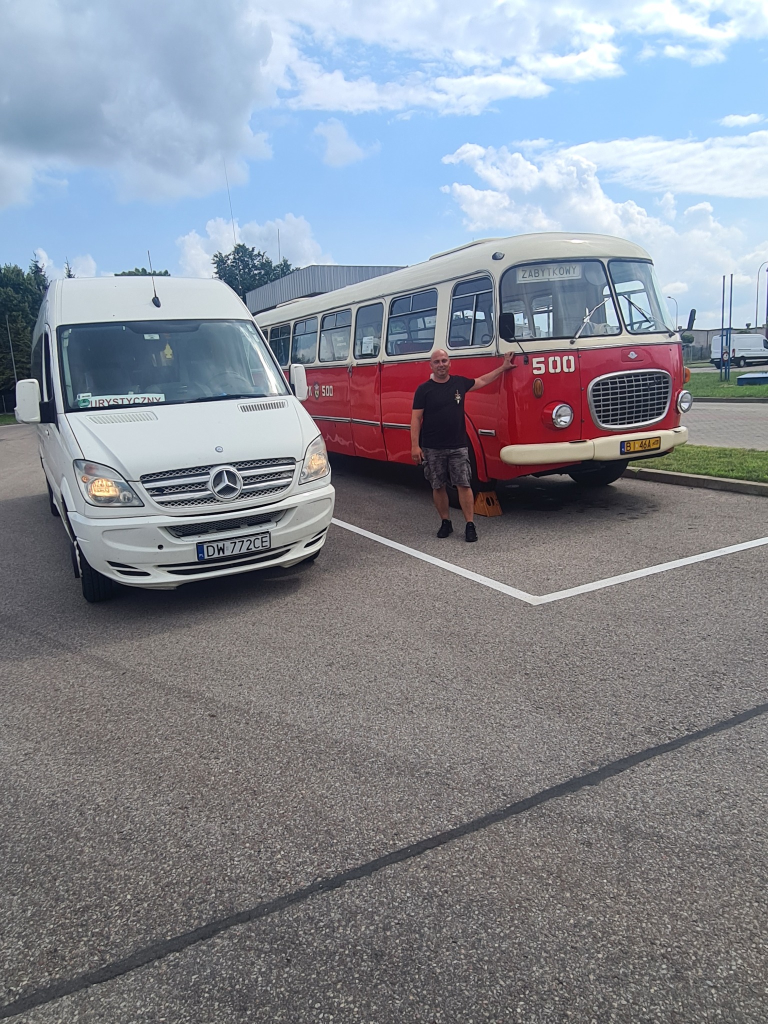 Biały Mercedes-Benz Sprinter z napisem 'Turystyczny' za szybą, obok czerwono-białego zabytkowego autobusu '500' z mężczyzną opierającym się o niego, zaparkowane na parkingu z wyznaczonymi miejscami.