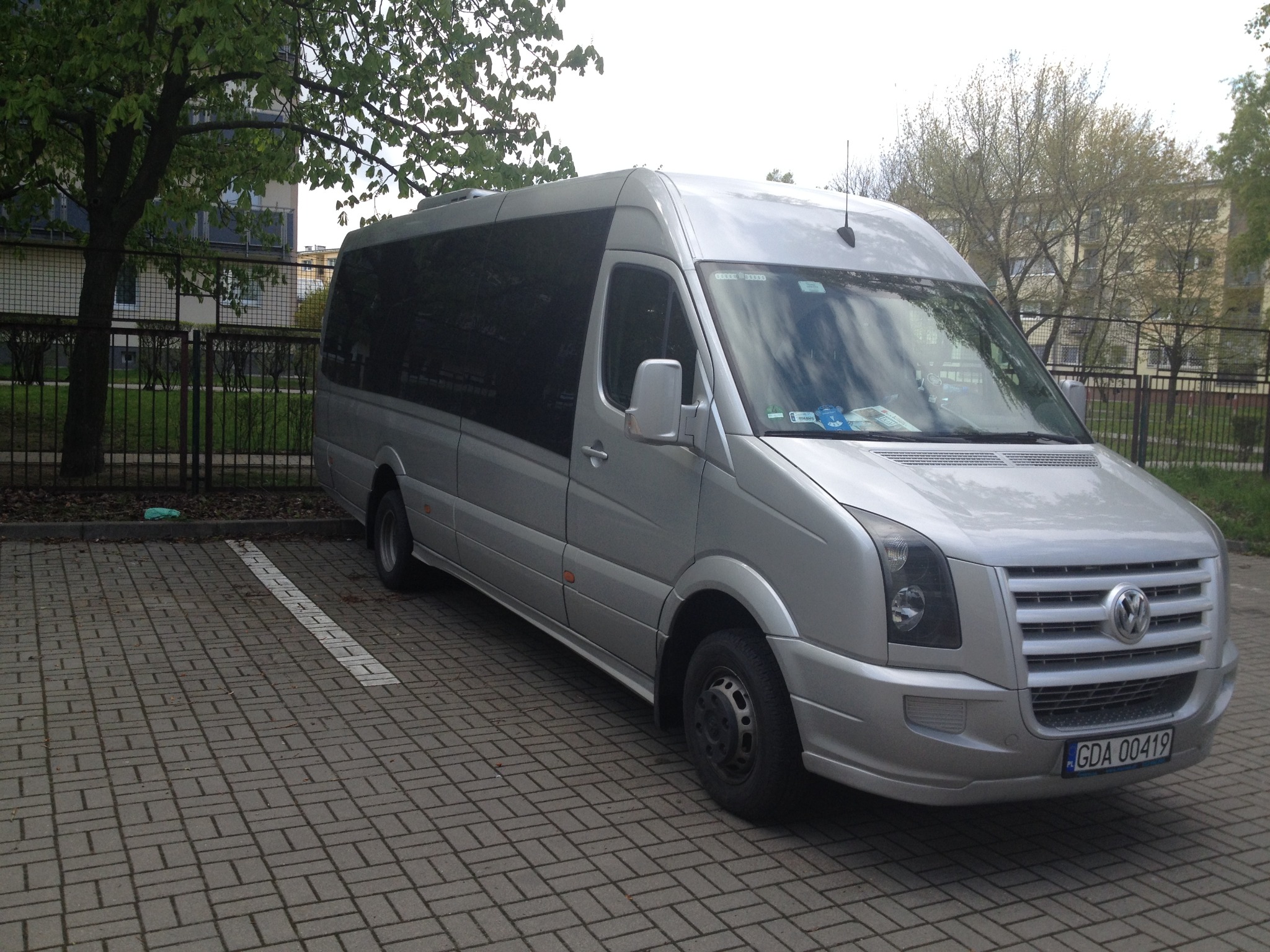 Srebrny bus Volkswagen Crafter z przyciemnianymi szybami, zaparkowany na parkingu z kostki brukowej, widoczne drzewa i budynek w tle.