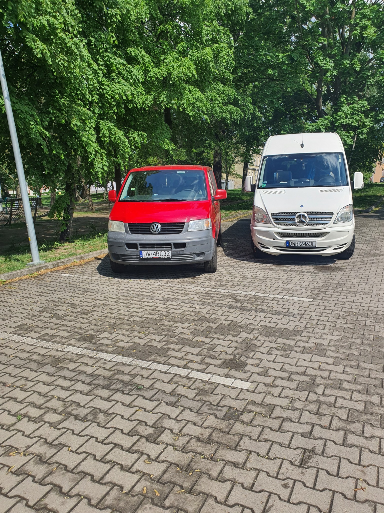 Dwa busy, czerwony Volkswagen Transporter i biały Mercedes-Benz Sprinter, zaparkowane obok siebie na parkingu wyłożonym kostką brukową, z drzewami w tle.