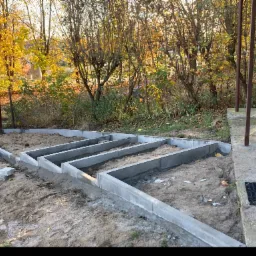 Betonowe fundamenty pod budowę, widok na jesienne drzewa w tle, częściowo wykopany teren, przygotowanie do dalszych prac budowlanych.