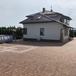 Rozległy podjazd z kostki brukowej w odcieniach brązu, z dekoracyjnym wzorem obramowania w kolorze białym, prowadzący do nowoczesnego domu z ciemnobrązowym dachem i balkonem; z boku widoczny basen...