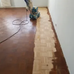 Parkiet w trakcie renowacji: połowa powierzchni wycyklinowana, ukazująca jasne drewno, druga połowa w oryginalnym, ciemniejszym odcieniu. Widoczna maszyna do cyklinowania z podłączonym kablem.