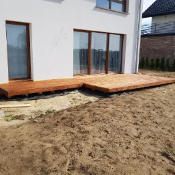Nowo wybudowany taras z drewna egzotycznego przylegający do białego domu z dużymi oknami, widoczne czarne podłoże pod konstrukcją tarasu i przygotowany teren wokół.