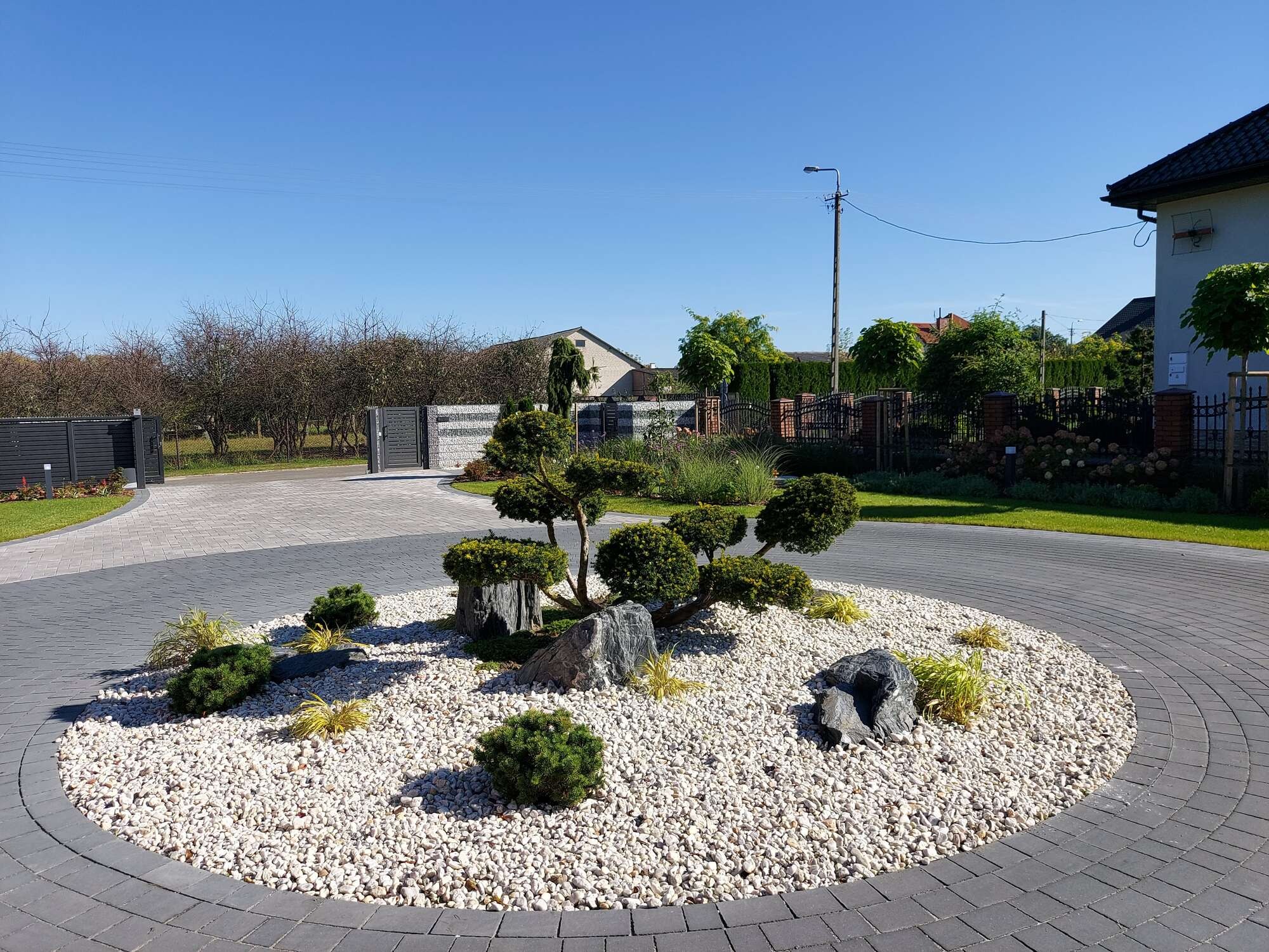 Okrągły klomb z białych kamieni, ozdobnymi drzewami i skałkami, otoczony kostką brukową w kolorze grafitowym, na tle zadbanego ogrodu z domami.