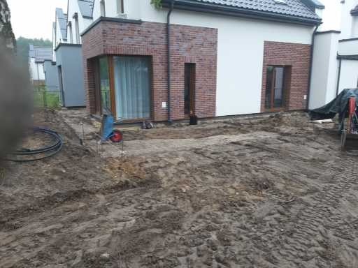 Przygotowany teren pod budowę domu jednorodzinnego, widoczne ślady po koparce, wyznaczone granice przyszłego trawnika, w tle budynek z ceglaną elewacją.