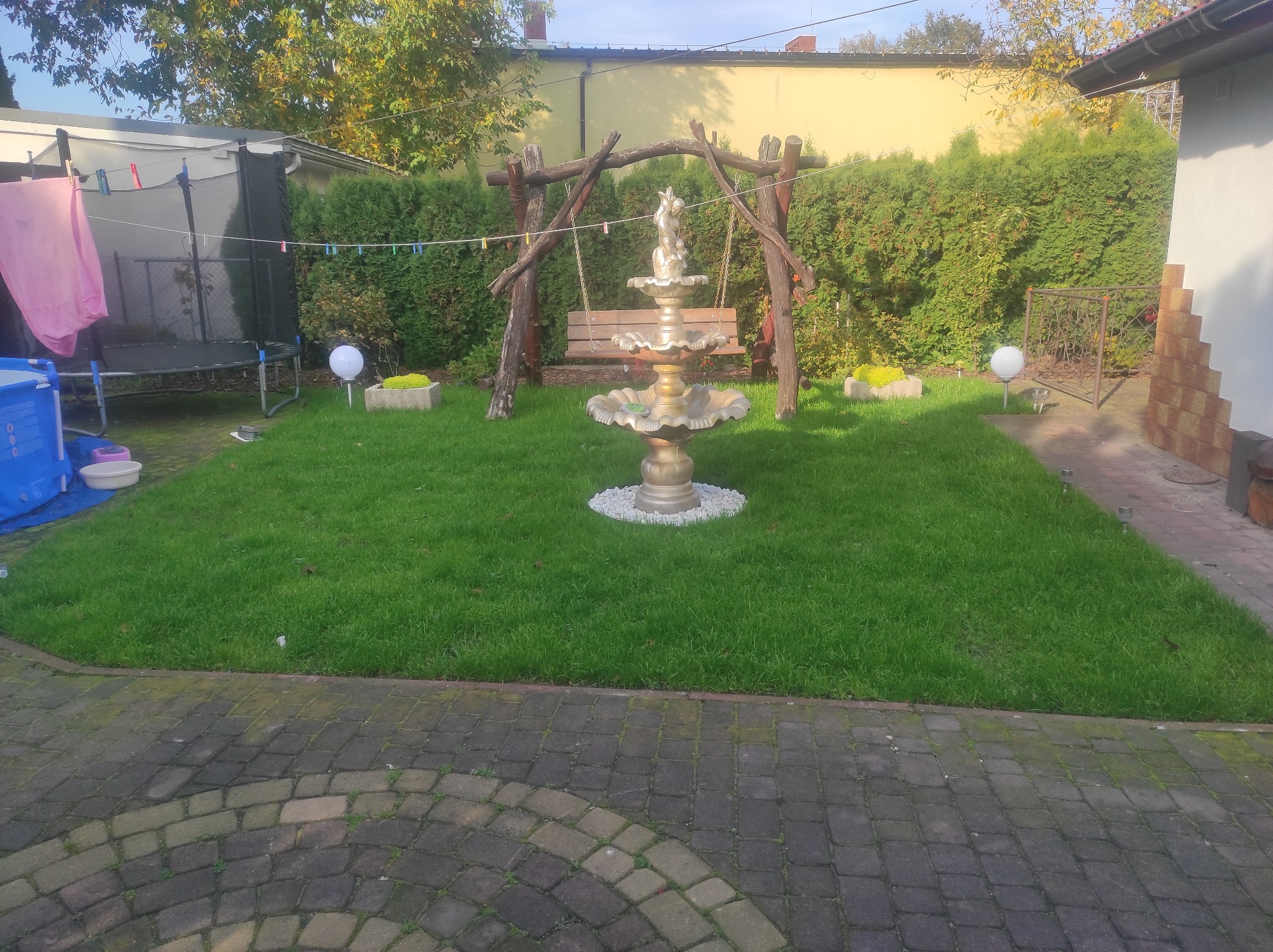 Ogród z fontanną w stylu rustykalnym i huśtawką z bali, trawnik, kostka brukowa, sznur z praniem w tle.