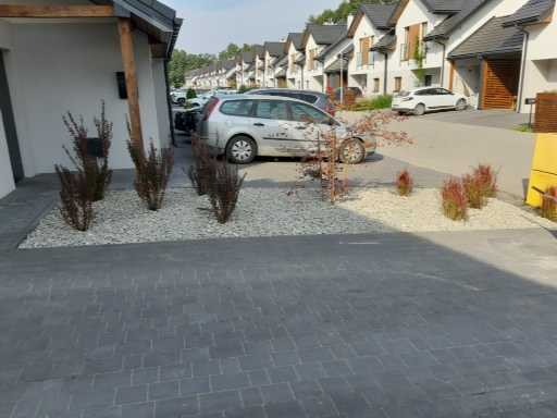 Nowoczesny podjazd wyłożony szarą kostką brukową, z białym żwirem i nasadzeniami ozdobnych traw przed szeregiem domów jednorodzinnych.