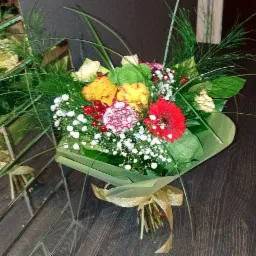 Ułożony bukiet z czerwonych, żółtych i białych kwiatów, owinięty w zielony papier i przewiązany złotą wstążką, leżący na ciemnym, drewnianym blacie obok lustrzanej powierzchni.