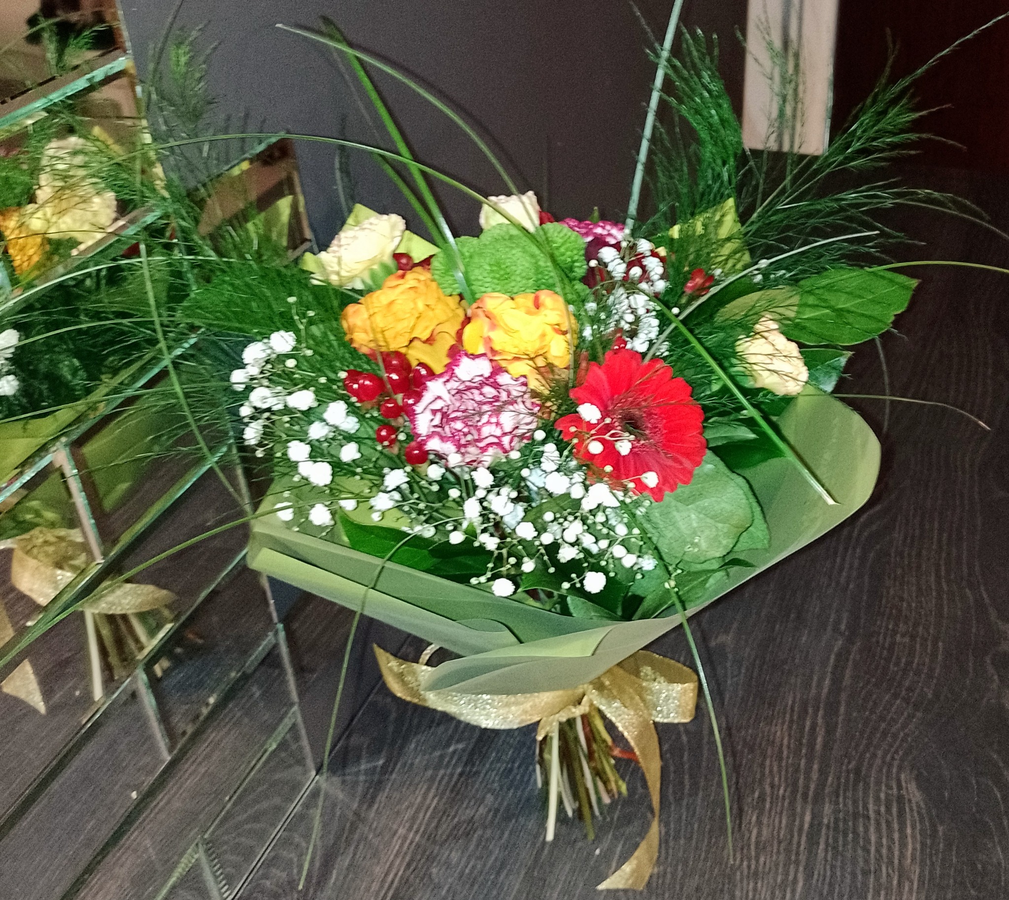 Ułożony bukiet z czerwonych, żółtych i białych kwiatów, owinięty w zielony papier i przewiązany złotą wstążką, leżący na ciemnym, drewnianym blacie obok lustrzanej powierzchni.
