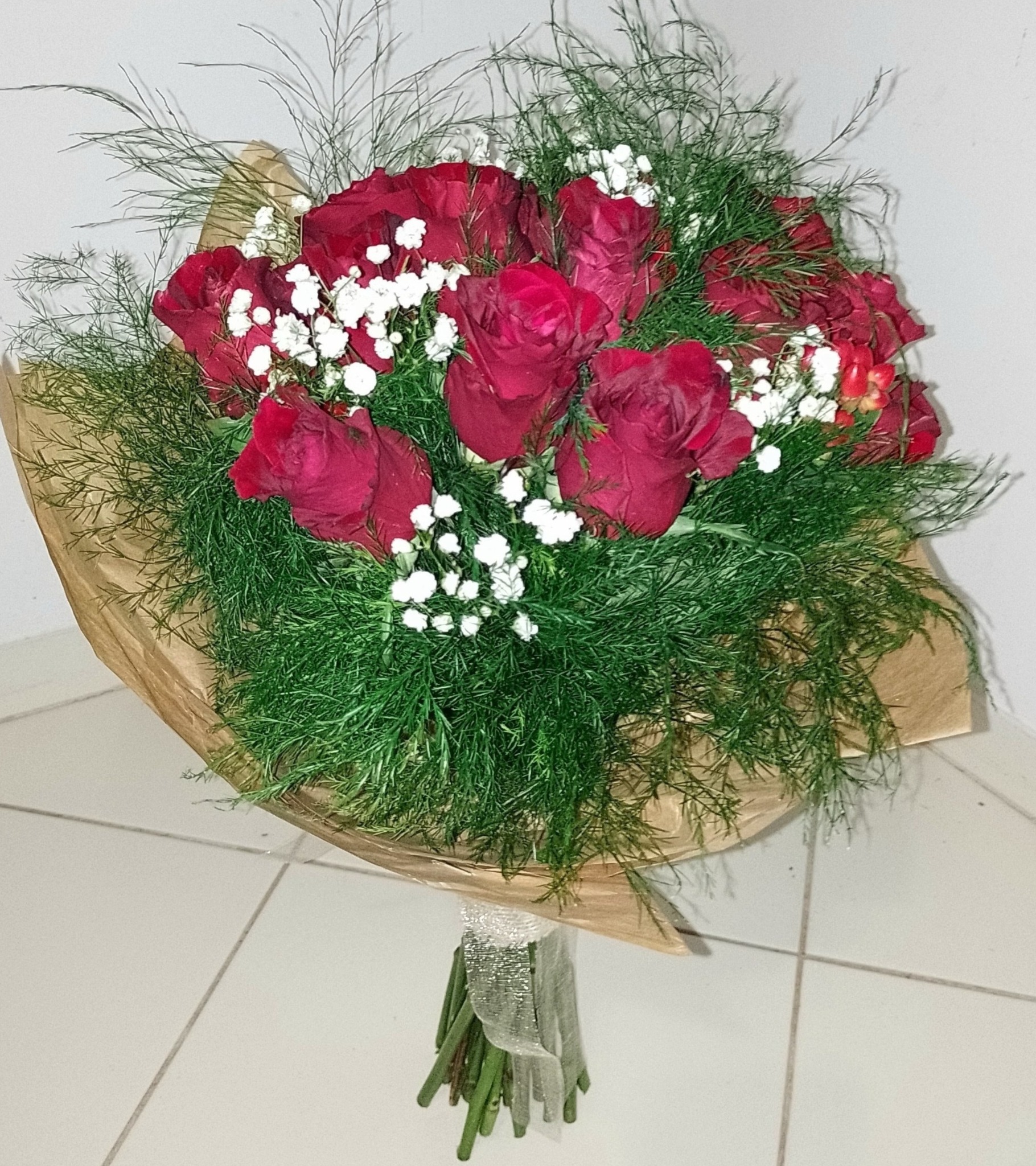 Bukiet czerwonych róż z gipsówką i zielonymi dodatkami, owinięty w brązowy papier i przewiązany srebrną wstążką, leżący na białych płytkach podłogowych.