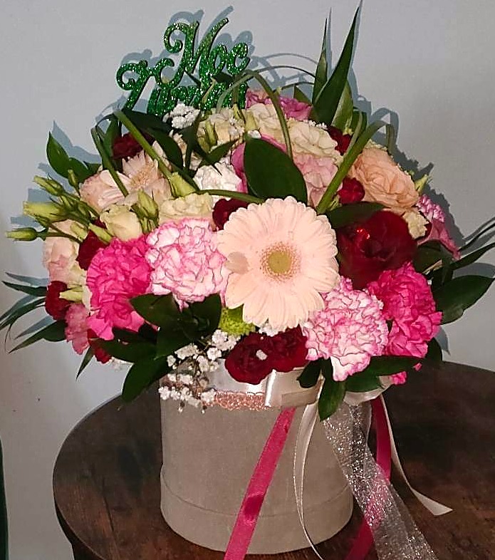 Kompozycja kwiatowa w szarym pudełku z różowymi i srebrnymi wstążkami, zawierająca gerbery, goździki, róże i zielony topper z napisem.