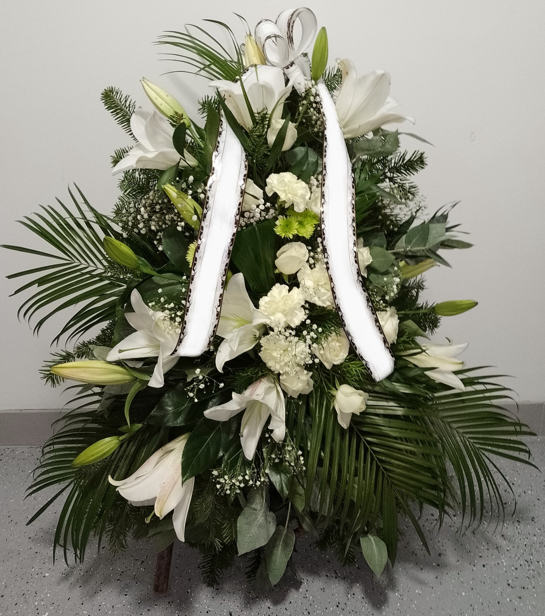 Biała kompozycja kwiatowa z lilii, goździków i zielonych liści palmowych, udekorowana białymi wstążkami z ciemnym obramowaniem, na tle szarej ściany i podłogi.