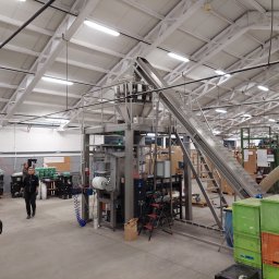 Marten - Wnętrze hali produkcyjnej z maszyną do automatycznego pakowania, widoczny podajnik taśmowy, stosy skrzynek i pracownik w tle.