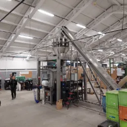 Wnętrze hali produkcyjnej z maszyną do automatycznego pakowania, widoczny podajnik taśmowy, stosy skrzynek i pracownik w tle.