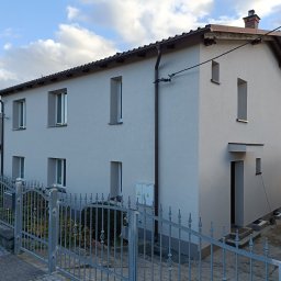 Dawid Kozikowski LUX - Dwukondygnacyjny dom jednorodzinny z nową, jasnoszarą elewacją, widoczny zza metalowego ogrodzenia z ozdobnymi elementami.