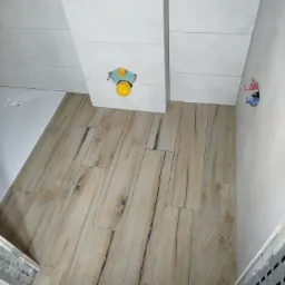 Wnętrze łazienki w trakcie wykańczania: ułożone płytki imitujące drewno na podłodze, białe płytki na ścianach, widoczne przyłącza wodne i odpływ prysznicowy.