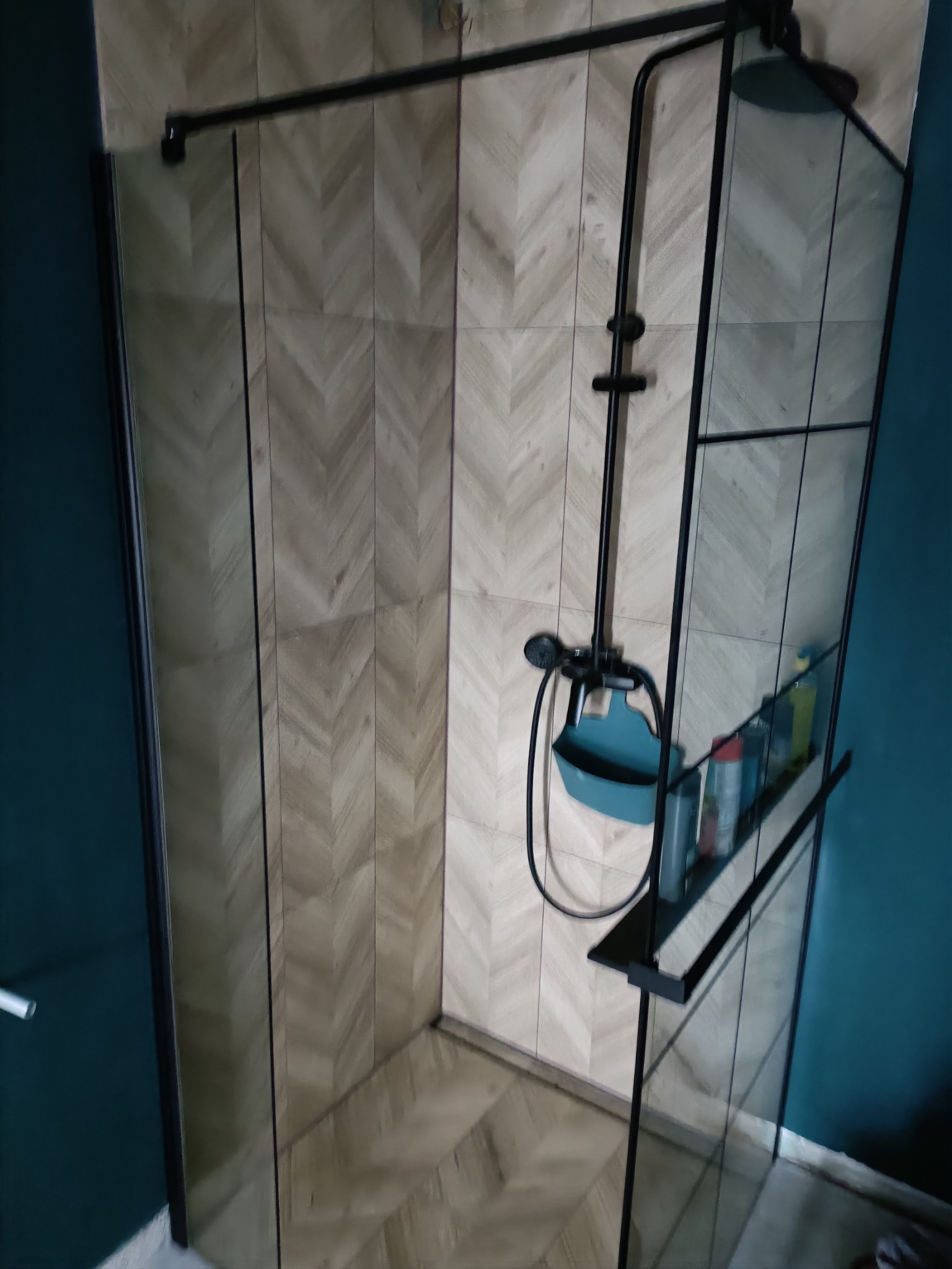 Kabina prysznicowa z czarnymi profilami i półką na kosmetyki, ściana wyłożona płytkami w jodełkę, widoczny fragment ściany pomalowanej na ciemny turkus.