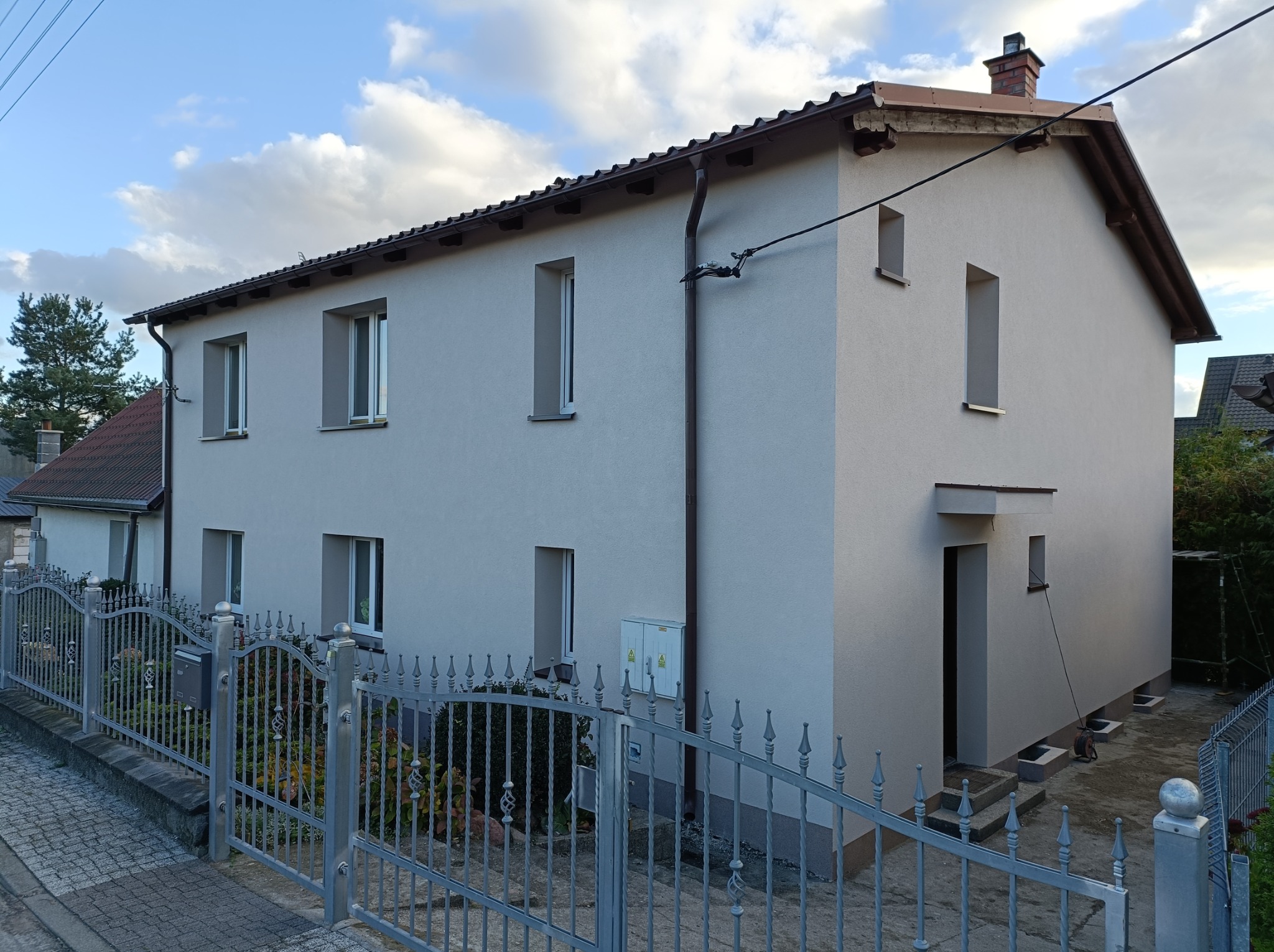 Dwukondygnacyjny dom jednorodzinny z nową, jasnoszarą elewacją, widoczny zza metalowego ogrodzenia z ozdobnymi elementami.