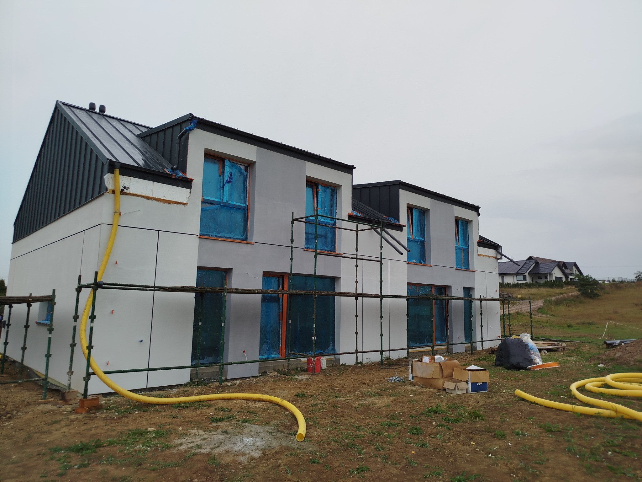 Nowoczesny dom w trakcie prac wykończeniowych elewacji, okna zabezpieczone folią, widoczne rusztowania i żółty wąż na ziemi.