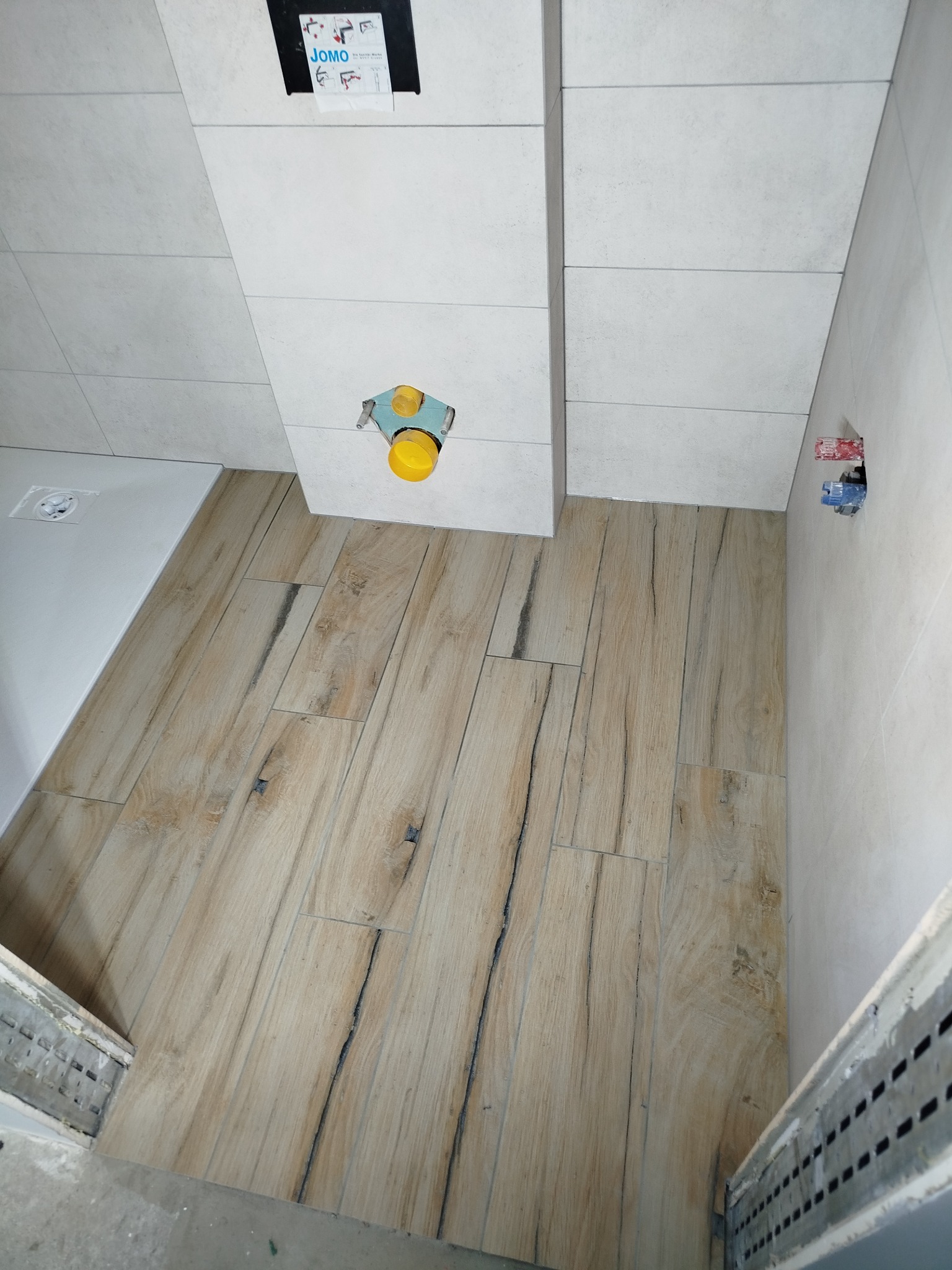 Wnętrze łazienki w trakcie wykańczania: ułożone płytki imitujące drewno na podłodze, białe płytki na ścianach, widoczne przyłącza wodne i odpływ prysznicowy.