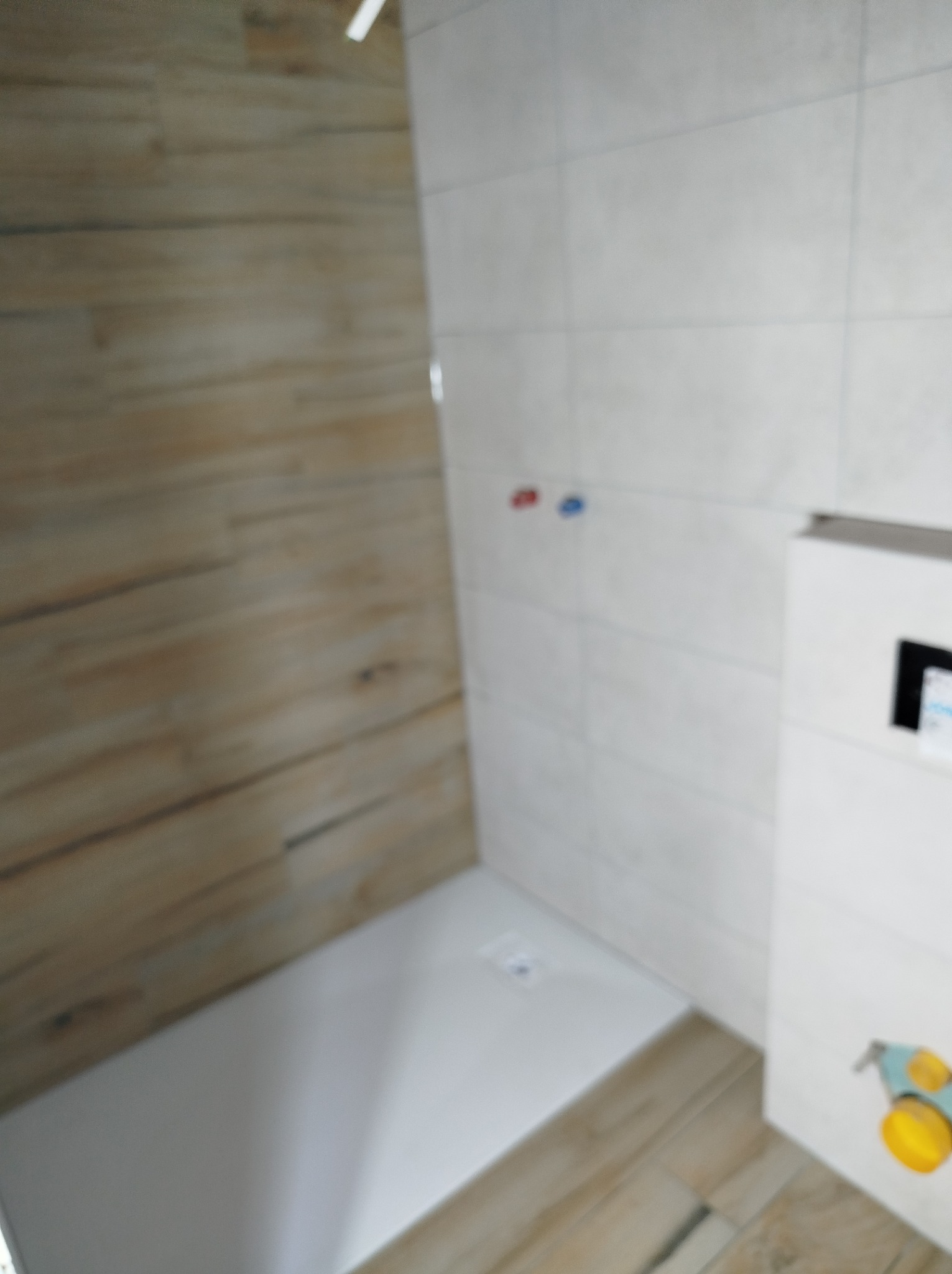 Nowoczesna kabina prysznicowa z drewnopodobnymi płytkami na ścianie i białymi płytkami na pozostałych ścianach, widoczne baterie i odpływ liniowy.