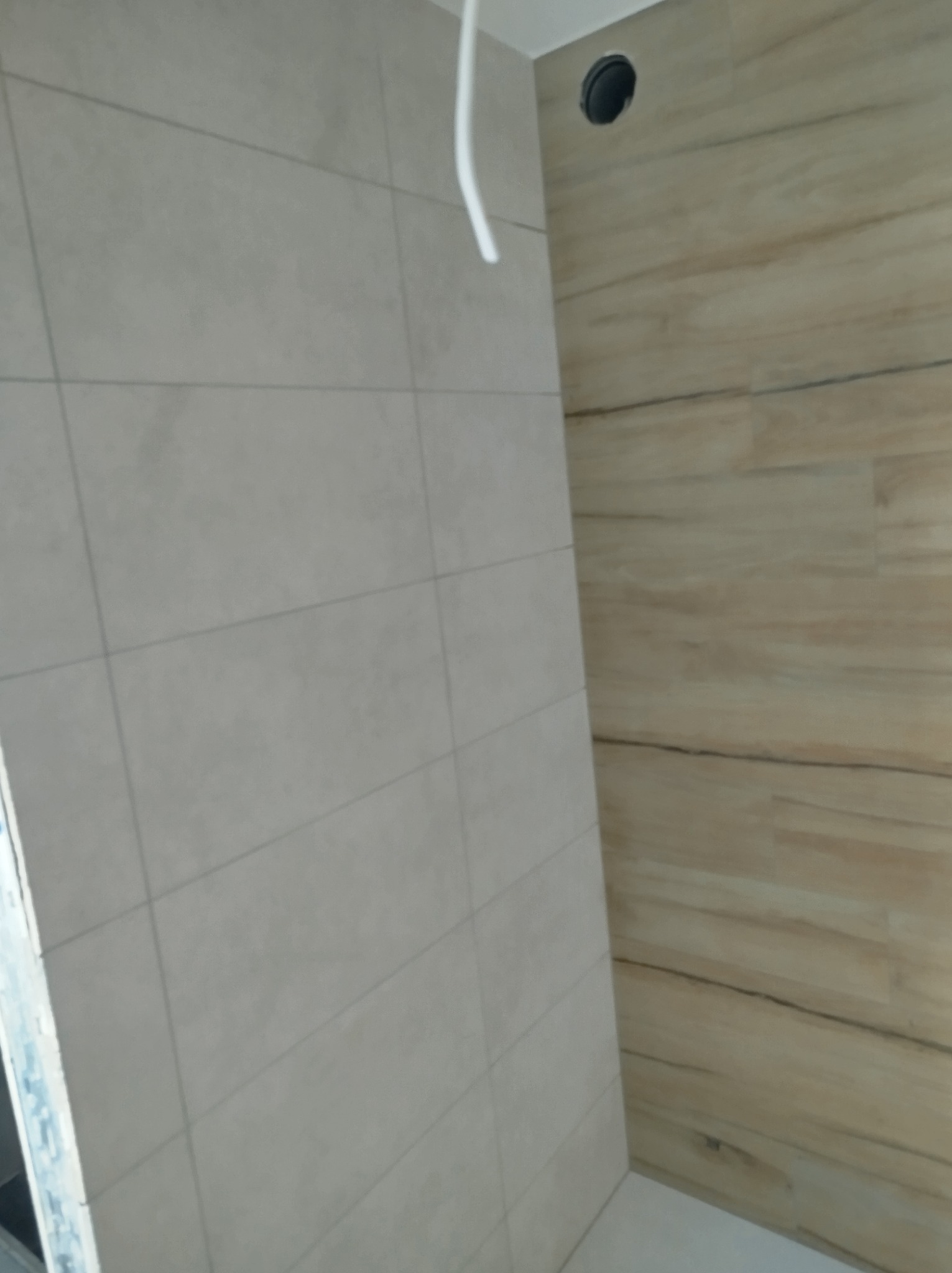 Fragment łazienki z wykończonymi ścianami: jedna wyłożona jasnymi, kwadratowymi płytkami, druga panelami imitującymi drewno; widoczny kabel elektryczny i otwór wentylacyjny w suficie.