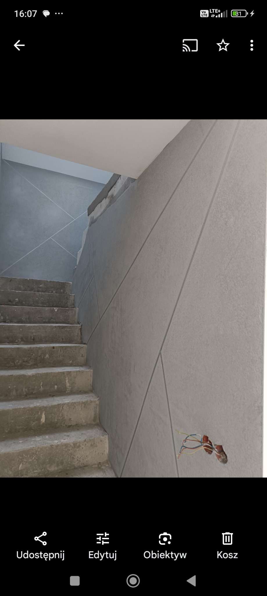 Ściana z szarej gładzi z dekoracyjnymi liniami i wystającymi kablami elektrycznymi obok surowych betonowych schodów w trakcie remontu.