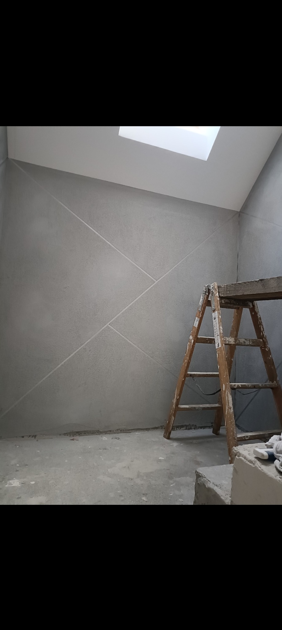 Ściana w szarym kolorze z geometrycznymi wzorami, drewniana drabina oparta o ścianę, okno dachowe w suficie, podłoga w trakcie prac wykończeniowych.