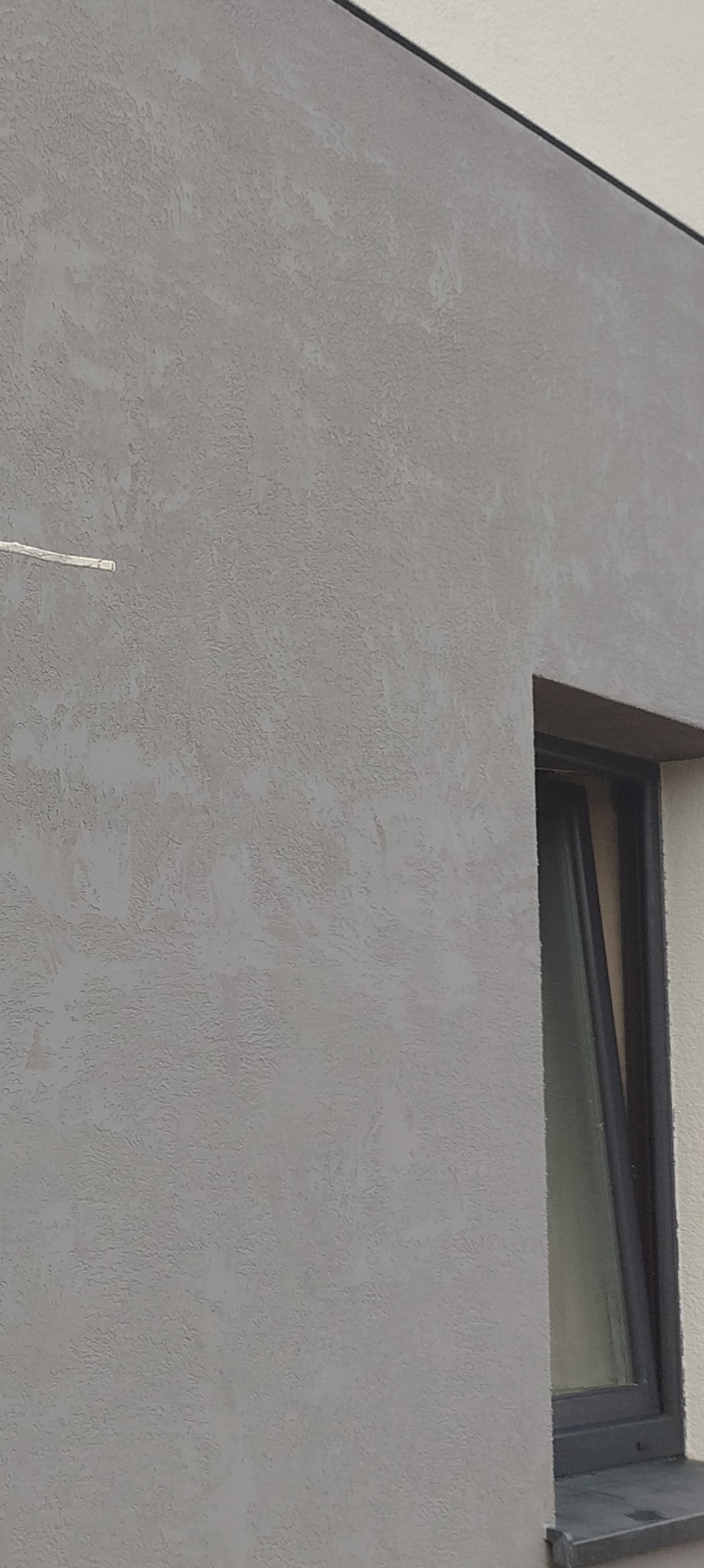 Szara, nierówna faktura tynku mineralnego na elewacji budynku, z widocznym fragmentem okna z ciemną ramą.