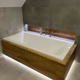 Nowoczesna wanna obudowana drewnem z podświetleniem LED, szara płytka na ścianie i podłodze, dekoracyjna mozaika miedziana nad wanną, bateria wannowa.