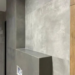 Szare, matowe płytki wielkoformatowe na ścianie w łazience z wnęką i przygotowanym otworem pod stelaż WC, widoczny fragment drewnopodobnej okładziny drzwi.