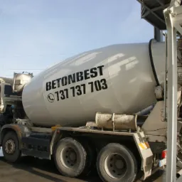 Ciężarówka z betonomieszarką marki BetonBest z numerem telefonu na bębnie, stojąca obok konstrukcji betoniarni w słoneczny dzień.