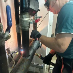 Spawacz w okularach ochronnych łączy rury instalacji grzewczej, widoczny płomień i iskry, w tle ściana z instalacjami.