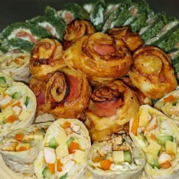 Dekoracyjna kompozycja przekąsek na drewnianej desce: roladki ze szpinakiem i łososiem, mini tortille z kurczakiem, serem i warzywami oraz słone paluszki z szynką.