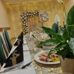 Uroczysty st&oacute;ł na osiemnaste urodziny z dekoracją balonową w odcieniach złota i srebra, eleganckimi czarnymi serwetkami ze złotym motywem, szklankami, talerzami z przekąskami i rośliną doniczkową...