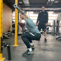 Mężczyzna wykonuje przysiad ze sztangą w siłowni, pod nadzorem trenera z napisem 'INSTRUKTOR' na bluzie.