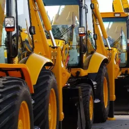 Rząd żółtych koparko-ładowarek zaparkowanych na placu, widoczne opony i kabiny z dokumentami 'Ready for Inspection'.
