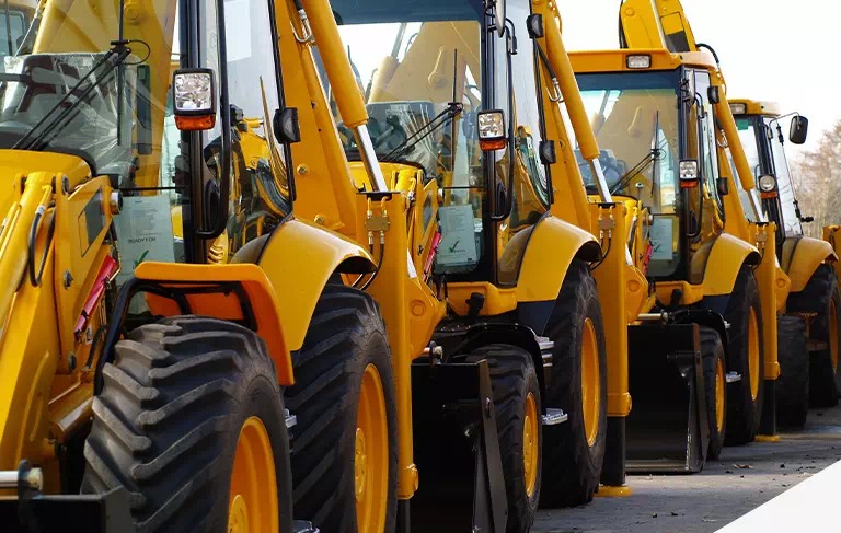 Rząd żółtych koparko-ładowarek zaparkowanych na placu, widoczne opony i kabiny z dokumentami 'Ready for Inspection'.