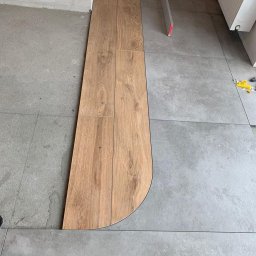 Pro-bud - Układanie paneli podłogowych z zaokrąglonym brzegiem, dopasowywanych do szarej, kwadratowej podłogi z płytek, z widoczną poziomicą i narzędziami w tle.