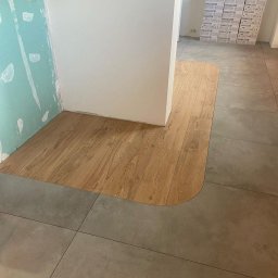 Pro-bud - Fragment podłogi z połączeniem płytek ceramicznych w odcieniach szarości i paneli drewnopodobnych, z zaokrąglonym przejściem między materiałami; w tle kartony z płytkami Marazzi.