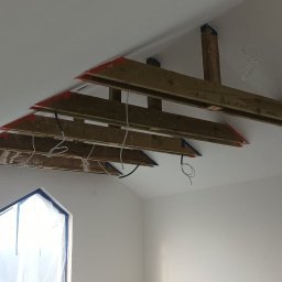 Pro-bud - Montaż drewnianej konstrukcji pod sufitem w nowym domu, widoczne kable i elementy instalacji elektrycznej, okno zabezpieczone folią.