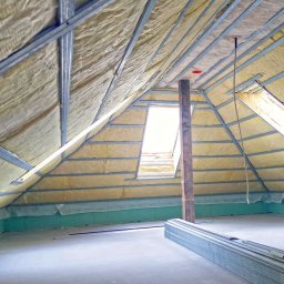 Pro-bud - Ocieplenie poddasza z oknem dachowym w Skierniewicach, widoczne metalowe profile i wełna mineralna, na podłodze stos metalowych elementów konstrukcyjnych.