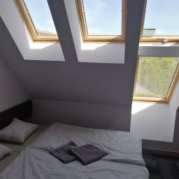 Pro-bud - Pokój na poddaszu z łóżkiem i trzema oknami dachowymi z widokiem na błękitne niebo i zieleń, ściany pomalowane na szaro.