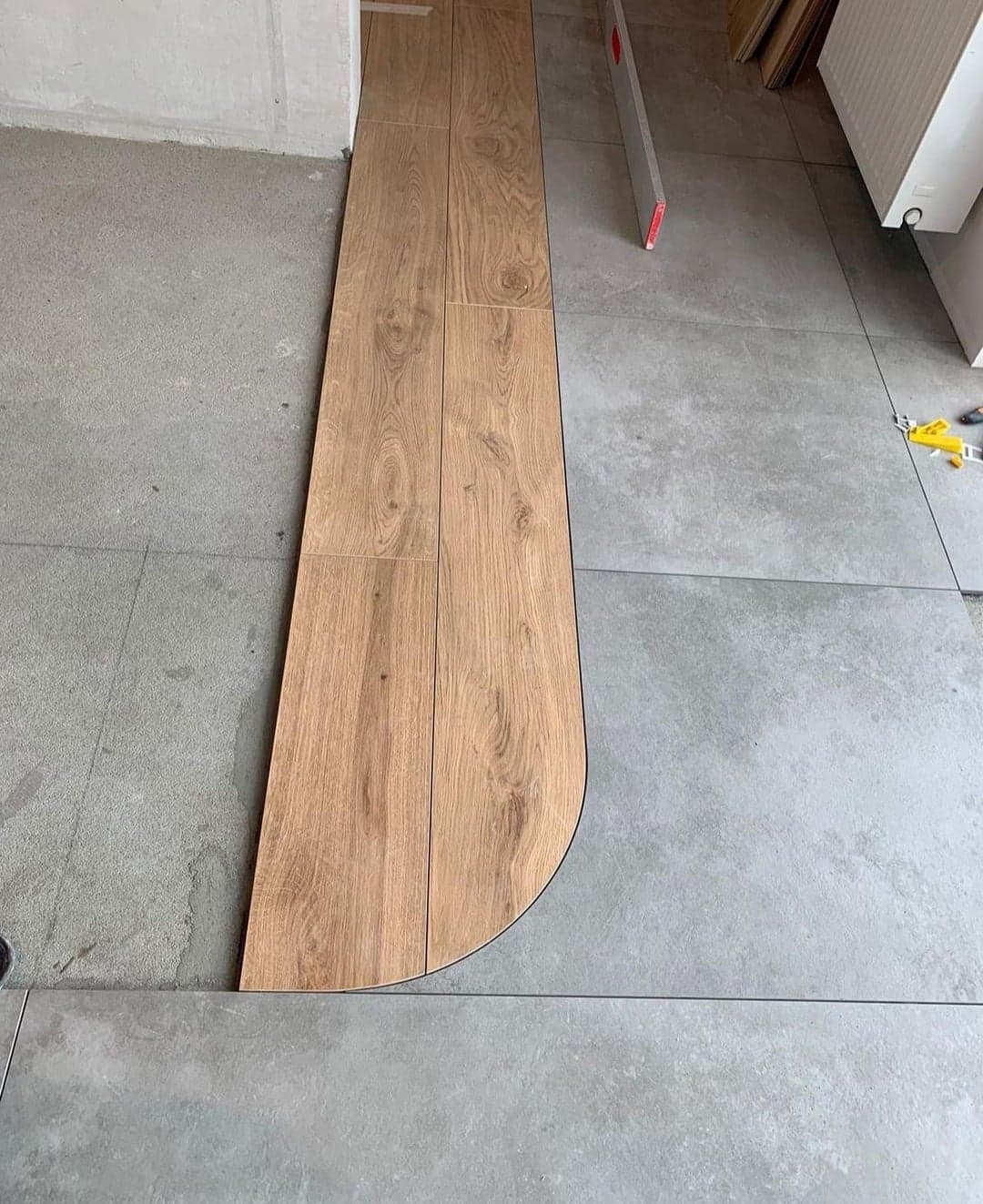 Układanie paneli podłogowych z zaokrąglonym brzegiem, dopasowywanych do szarej, kwadratowej podłogi z płytek, z widoczną poziomicą i narzędziami w tle.