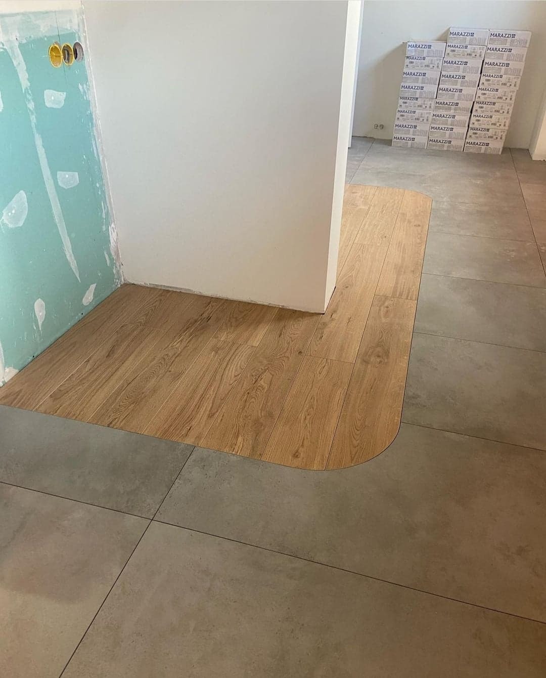 Fragment podłogi z połączeniem płytek ceramicznych w odcieniach szarości i paneli drewnopodobnych, z zaokrąglonym przejściem między materiałami; w tle kartony z płytkami Marazzi.