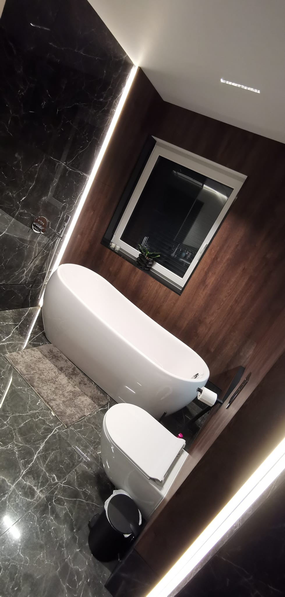 Nowoczesna łazienka z wolnostojącą wanną, toaletą i czarną marmurową okładziną ścian, oświetlona listwami LED wzdłuż krawędzi ścian.