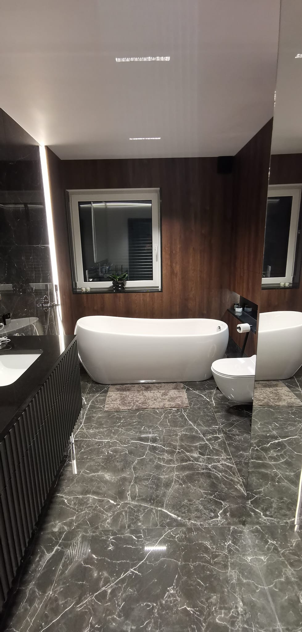 Nowoczesna łazienka z wolnostojącą białą wanną, podwieszaną toaletą, czarnymi marmurowymi płytkami podłogowymi i drewnianą okładziną ściany, oświetlona listwami LED.